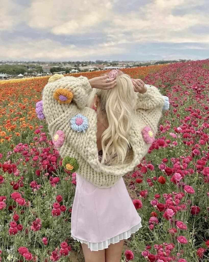 A woman with long blonde hair, wearing a chunky cream sweater with colorful flower patterns and a pale pink dress, stands in a vibrant field of blooming pink and red flowers under a cloudy sky.