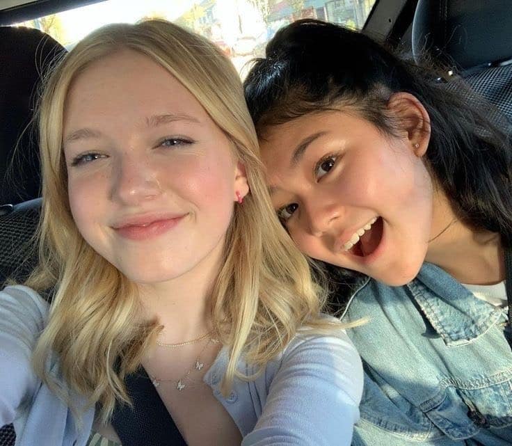 Two teenage girls, transitioning to high school, sit in a car smiling for a selfie. The blonde girl on the left wears a light blue top, while her friend on the right sports dark hair and a denim jacket—capturing memories of this exciting new chapter.