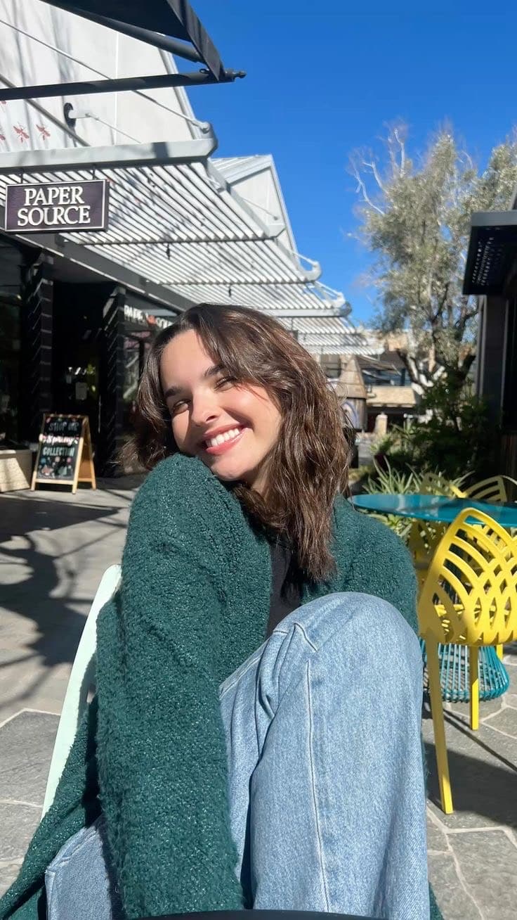 A young woman with wavy brown hair sits outside on a yellow chair, smiling brightly at the camera. She wears a green sweater and blue jeans, enjoying a sunny day near a Paper Source store while reflecting on her high school transition.