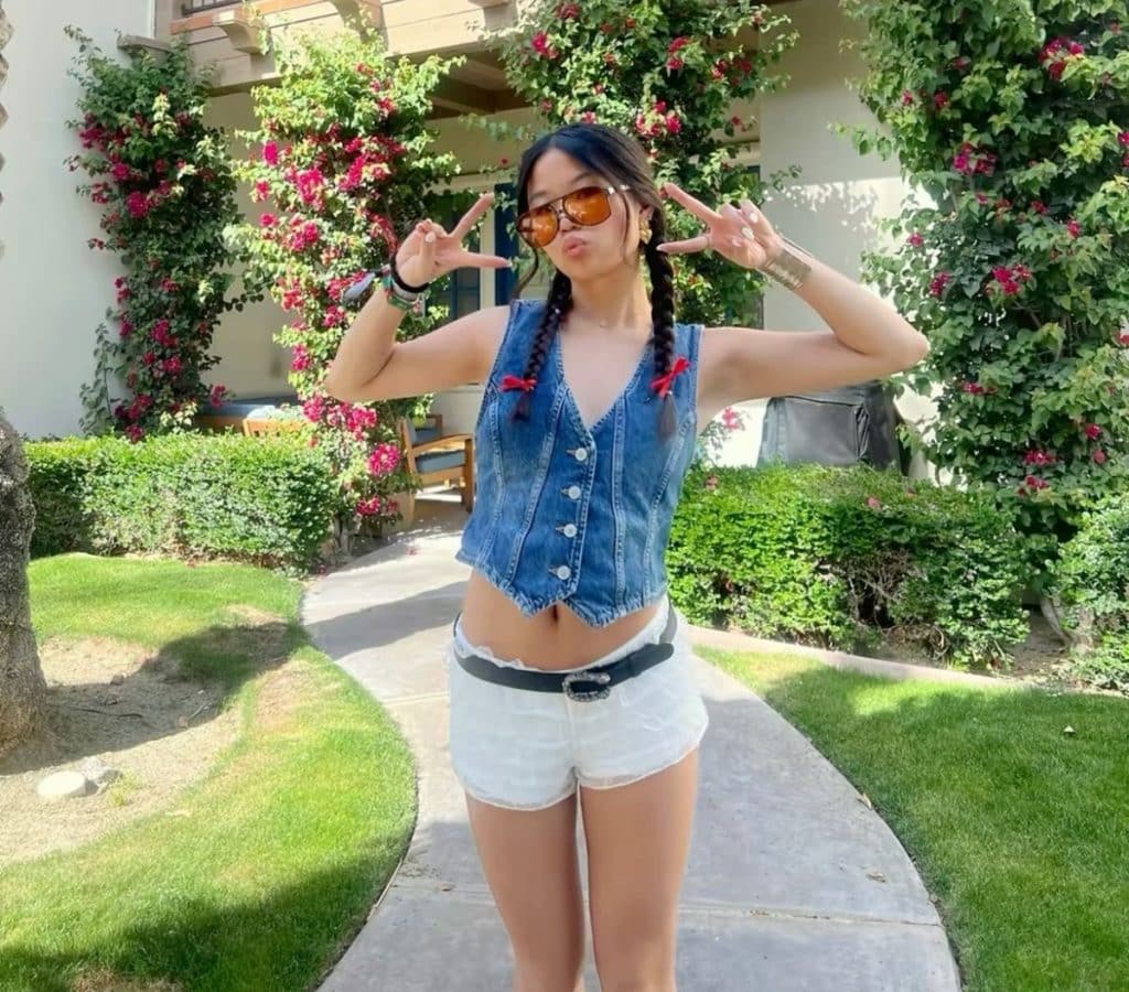 A woman with braided hair poses outdoors, making peace signs with both hands. Her playful style, much like the transition from middle school to high school, shines in her sunglasses, sleeveless denim top, and white shorts against lush greenery and flowers.