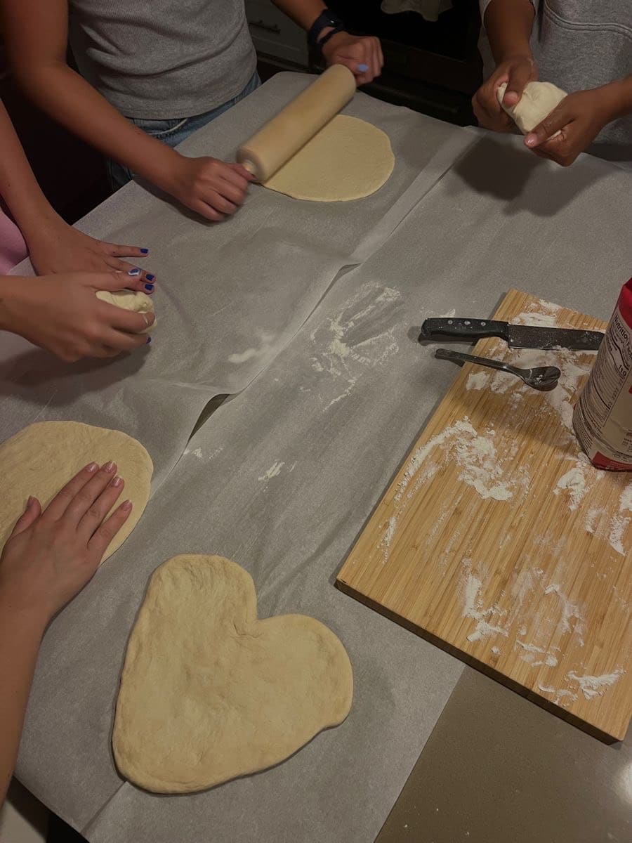 Four people shape pizza dough on parchment paper—one rolls with a pin, and one dough is heart-shaped. A wooden board with flour, utensils, and a canister sits nearby. Perfect inspiration for your next DIY pizza night!.