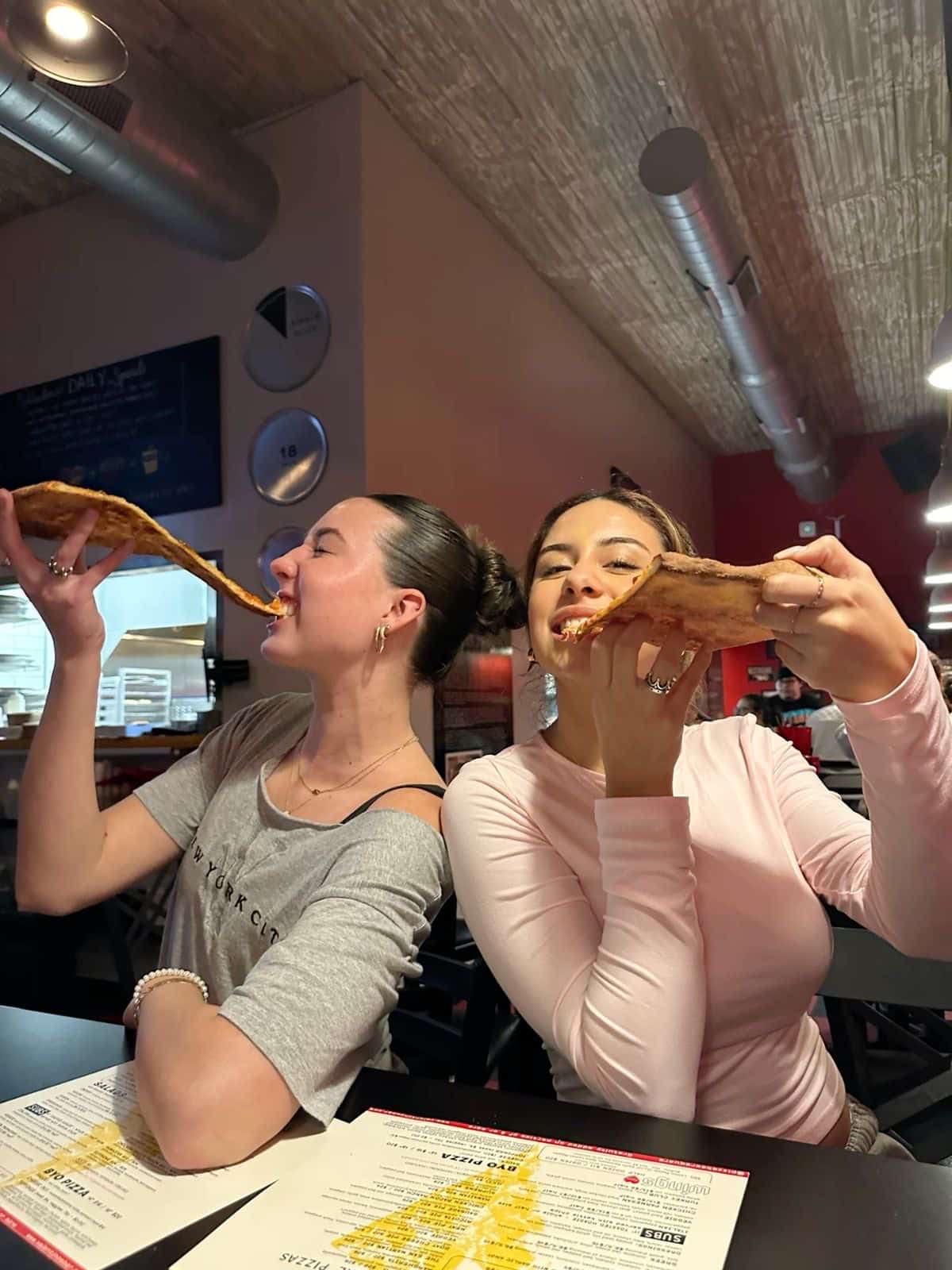 Two young women sit side by side in a cozy restaurant, smiling and holding up large slices of pizza. The warm lighting and menus on the table inspire fun pizza night ideas perfect for your own DIY pizza night at home.