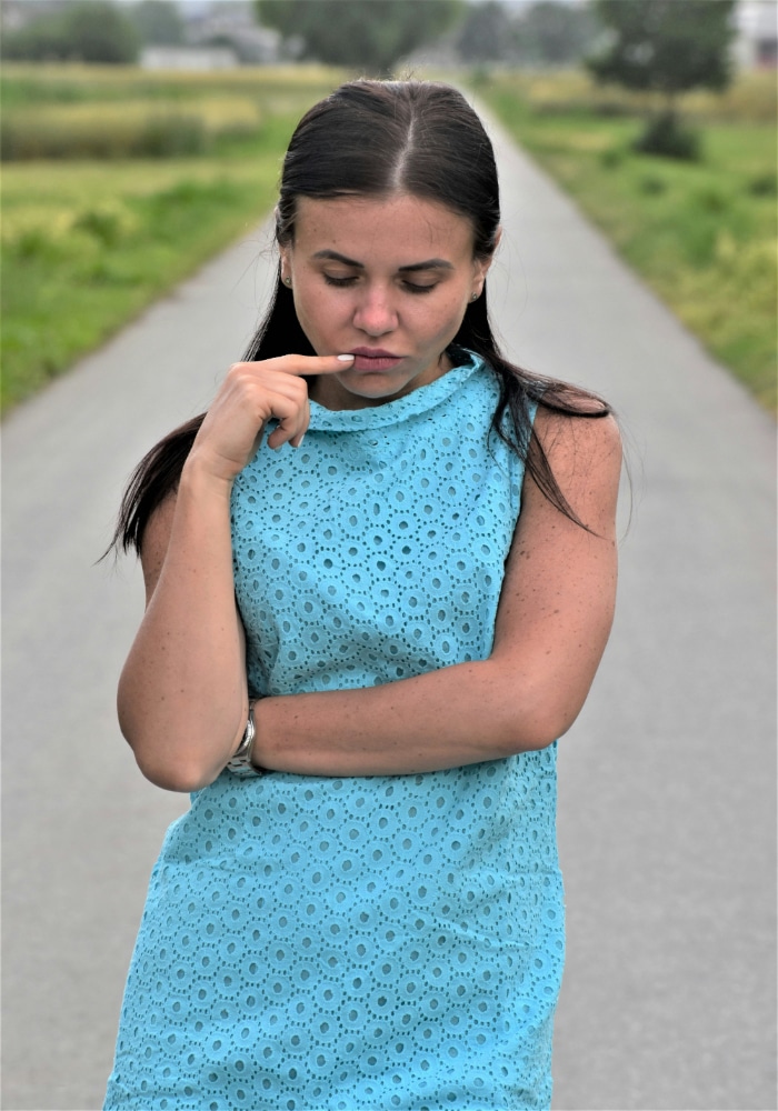 A woman in a sleeveless blue dress stands on a rural road with her arms crossed, looking down thoughtfully and touching her lip with one finger, appearing insecure in her relationship. Green fields and trees line the road in the background.