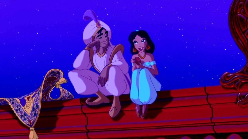 A young man in a turban and a woman in blue clothes sit together on a red rooftop at night, smiling at each other. A decorated magic carpet floats nearby against a starry sky, evoking the wonder of childhood flicks and unforgettable V-Day moments.