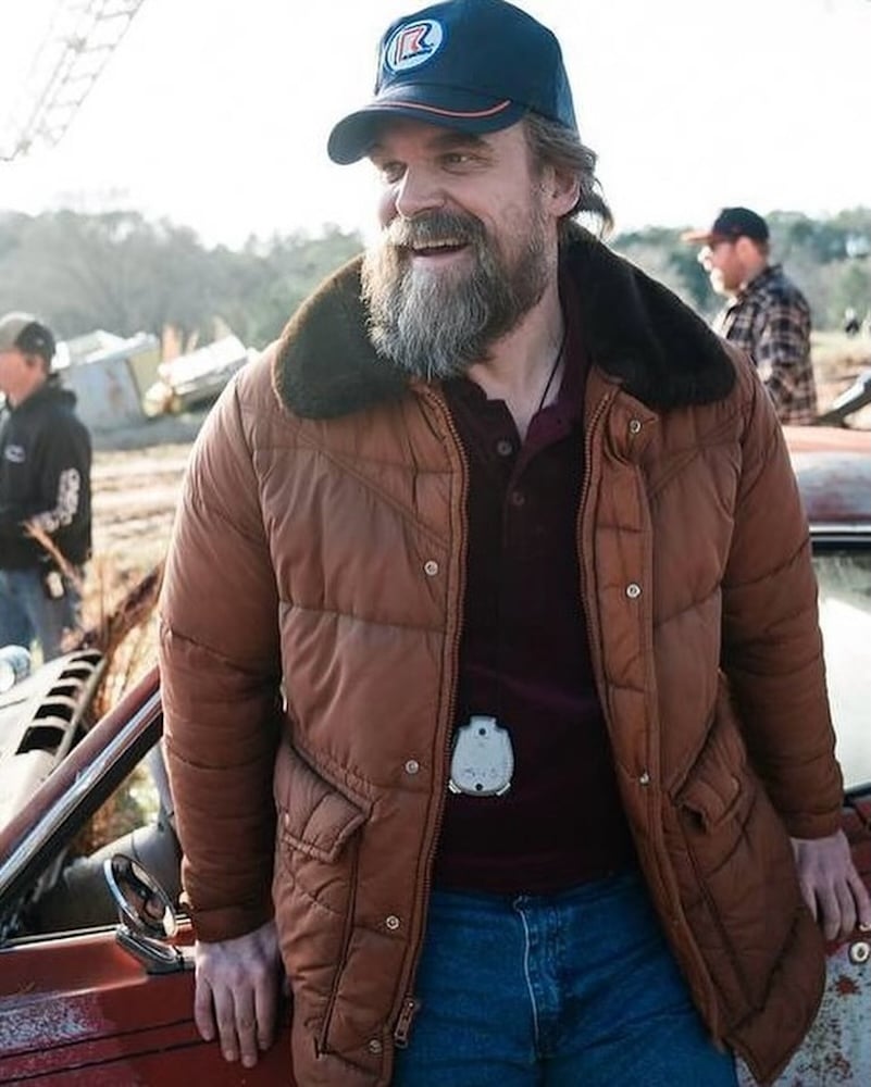 A bearded man in a brown winter jacket and blue cap stands smiling beside an old red car outdoors, resembling a Stranger Things character. He wears blue jeans with a police badge on his belt. Other people are visible in the background.