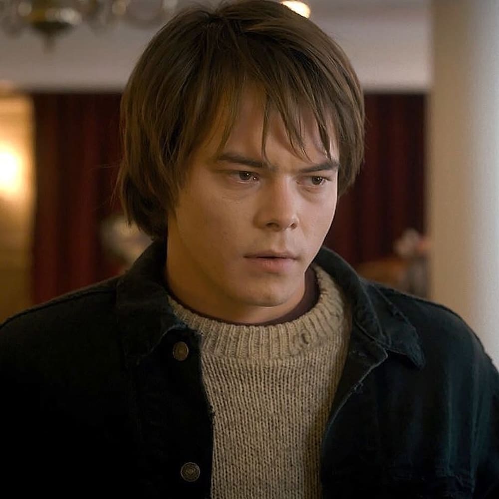 A young person with straight brown hair and a serious expression, reminiscent of a Stranger Things character, wears a cream sweater and dark jacket, standing indoors with blurred red curtains in the background.