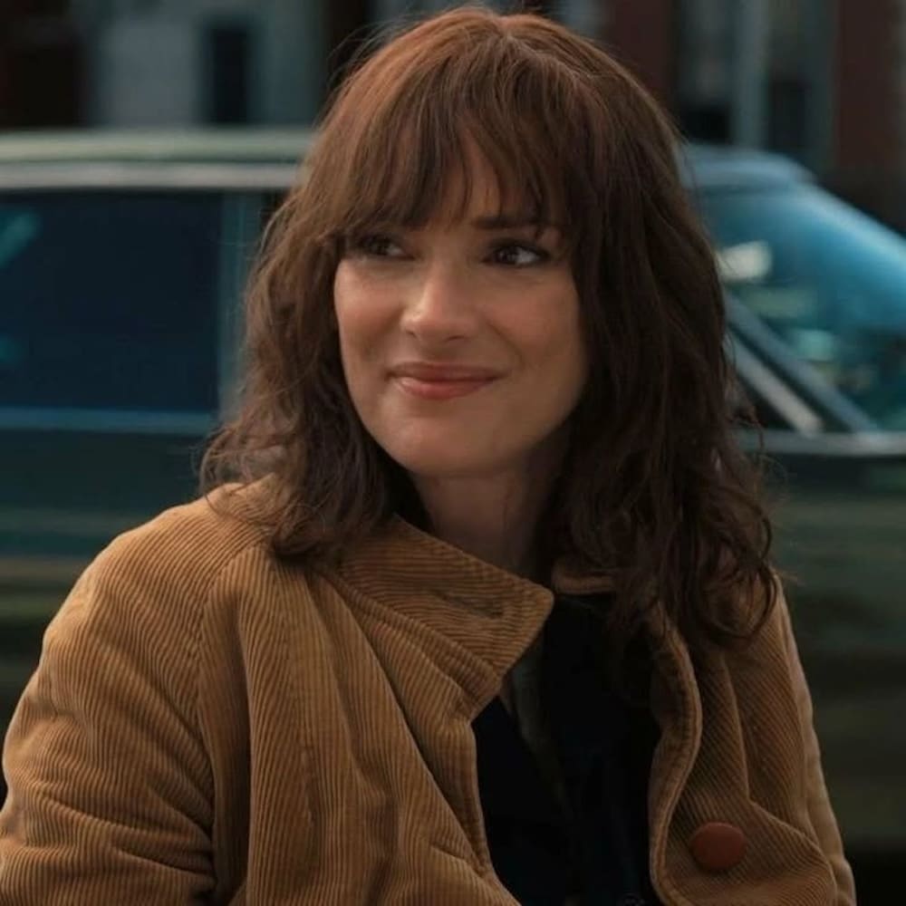 A woman with shoulder-length brown hair and bangs, resembling a character from Stranger Things, smiles in a tan corduroy jacket while sitting outdoors, with a blurred car in the background.