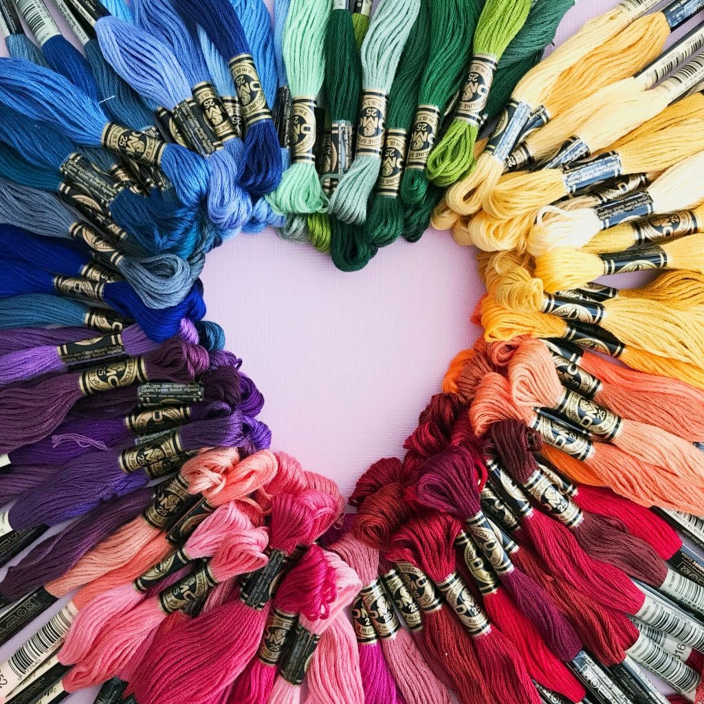 Skeins of embroidery floss in various colors are arranged in a heart shape, forming a vibrant rainbow gradient on a light pink background—a perfect inspiration to Sell Your Craft and showcase your handmade talent.