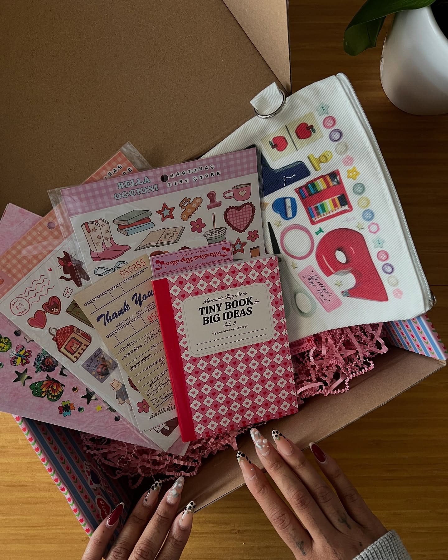 A person with decorated nails opens a box filled with pink shredded paper, stickers, a “Tiny Book of Big Ideas” notebook, a thank you note, and various stationery and craft items on a wooden table.