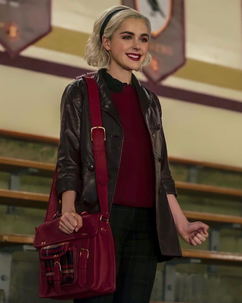 A young woman with blonde hair, wearing a dark leather jacket, red top, and carrying a red satchel, stands smiling in front of bleachers and wall banners inside a gymnasium.