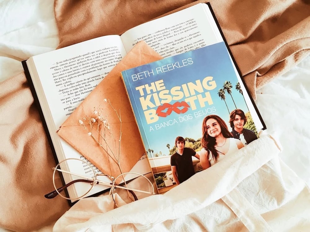 A copy of The Kissing Booth by Beth Reekles, one of the most popular Wattpad novels, rests on an open book beside a brown envelope, eyeglasses, and dried flowers, all set against a beige fabric background.