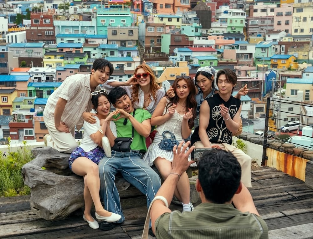 A group of seven young adults pose playfully for a photo outdoors, smiling and making finger heart gestures, with colorful hillside houses in the background—perfect for fans of XO, Kitty Season 3. One person sits in front, taking their picture.