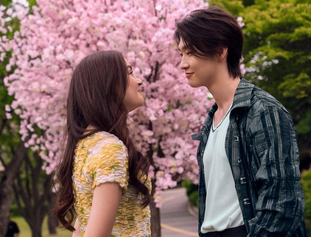 A young couple stands face to face, smiling at each other under blossoming pink cherry trees in a park. The scene radiates springtime joy—just like XO, Kitty Season 3 fans choose their favorite romantic moments.