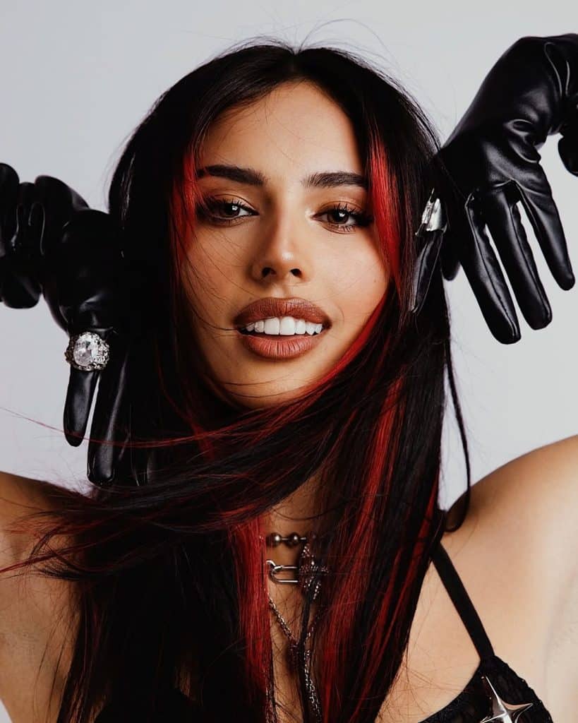 A woman with long dark hair and red streaks poses, wearing black gloves and jewelry. Her hands are near her face, and she is smiling slightly, with a confident expression, against a plain background.