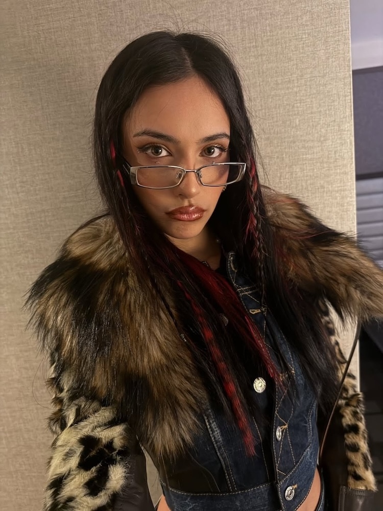 A woman with long dark hair and red streaks wears glasses, a faux fur-collared jacket, and a serious expression, posing in front of a textured beige background.