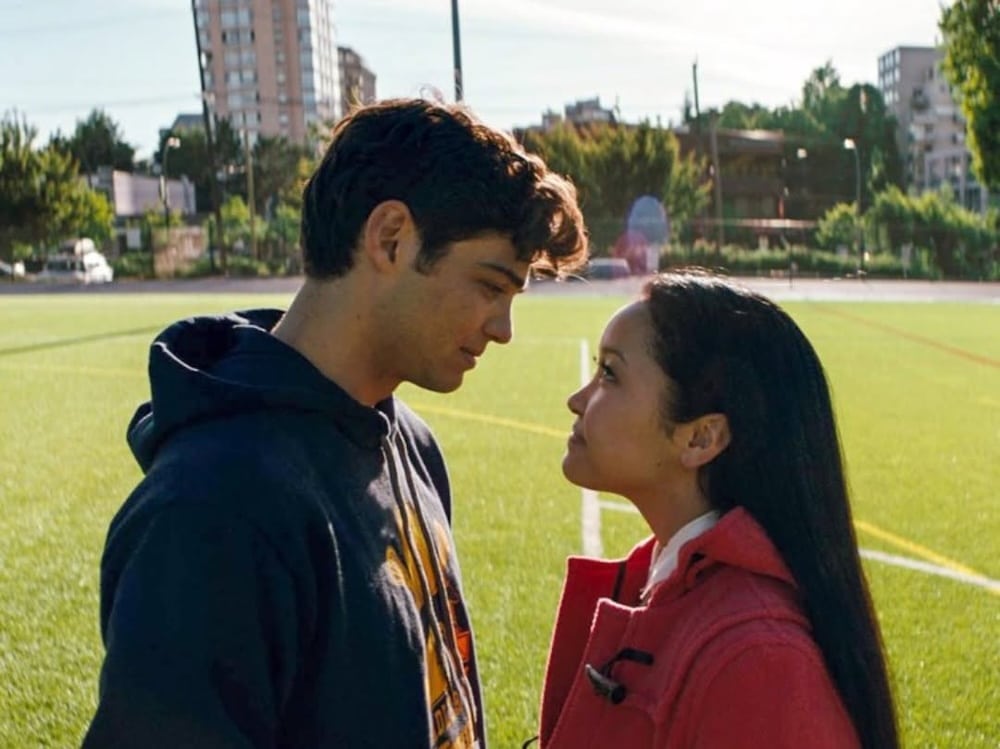 A young man and woman stand face to face on a sports field, gazing into each other's eyes like a scene from romantic comedies. The man wears a dark hoodie and the woman a red coat, with buildings and trees in the background.