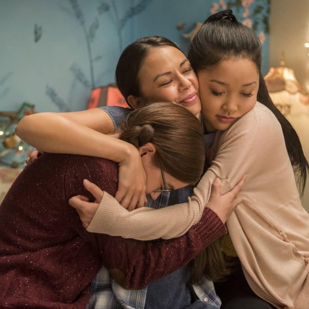 Three young women sit close together in a warm, comforting group hug, sharing an XO moment. One rests her head down while the others appear content, cozied up in a softly lit room reminiscent of Kitty Season 3’s heartfelt scenes.