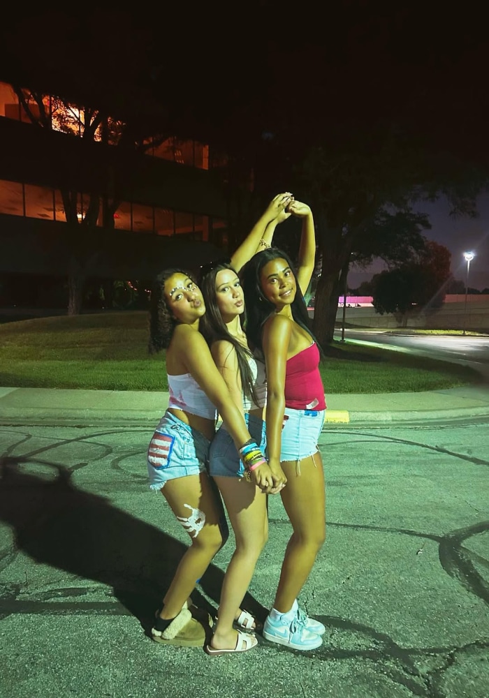 Three young women pose together at night in a parking lot, smiling and holding hands. They wear summer outfits with shorts and tank tops, and streetlights and a building glow in the background.