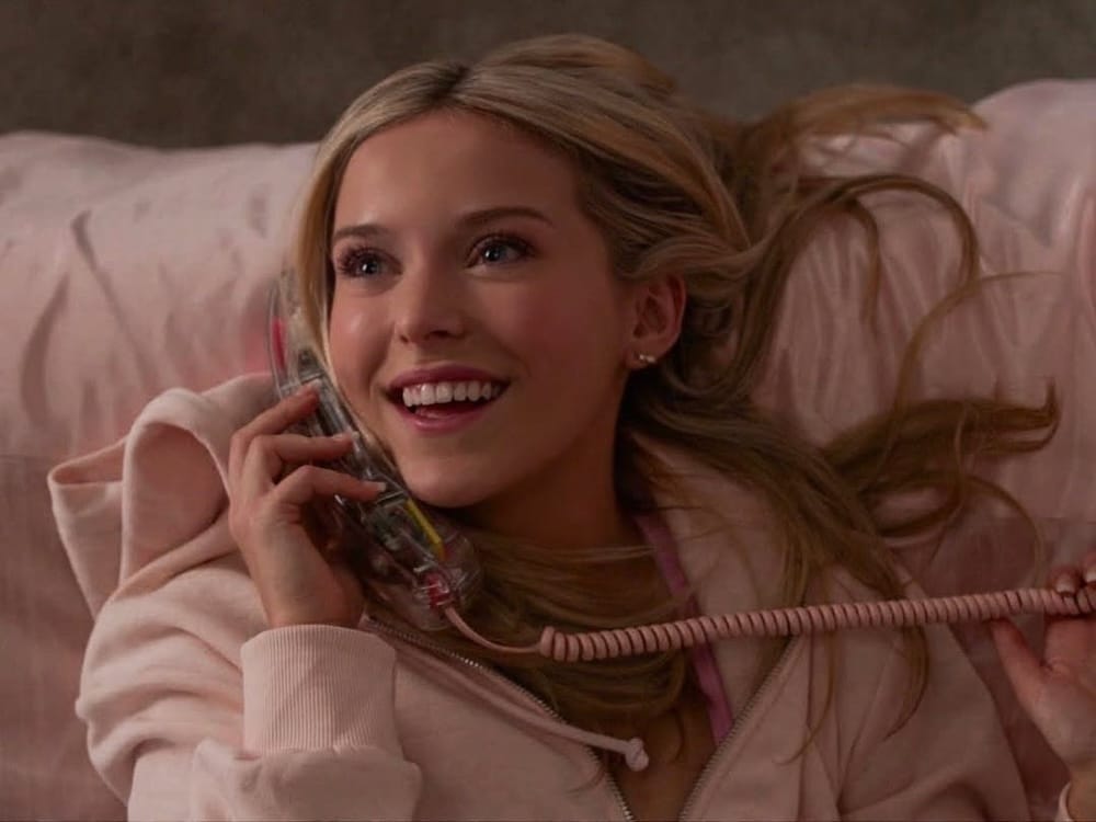 A young woman with long blonde hair, wearing a light pink hoodie, lies on a bed with pink bedding, smiling brightly while talking on a clear landline phone—a scene inspired by Elle from Legally Blonde’s Prequel Series.