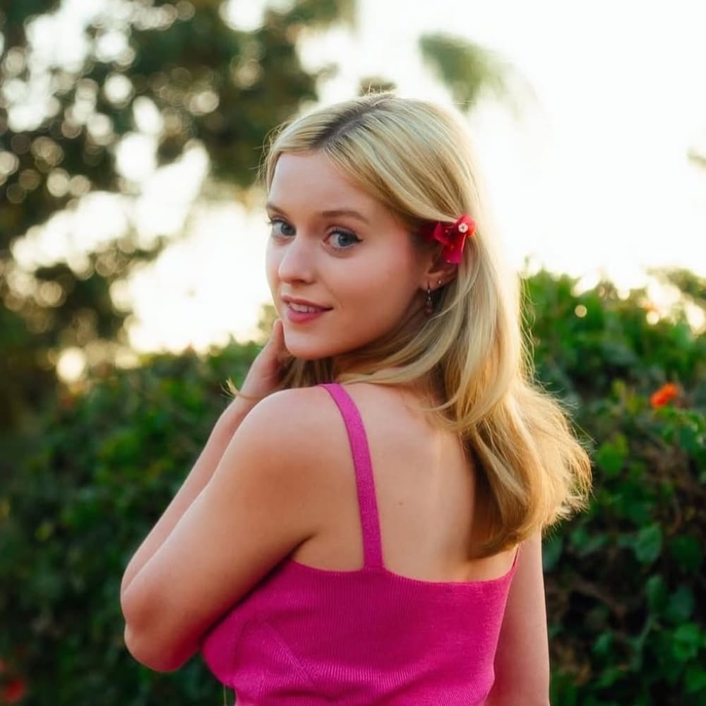 A young woman with blonde hair, reminiscent of Elle from the Legally Blonde series, wears a bright pink top and a red flower in her hair as she smiles over her shoulder, framed by greenery and soft sunlight.