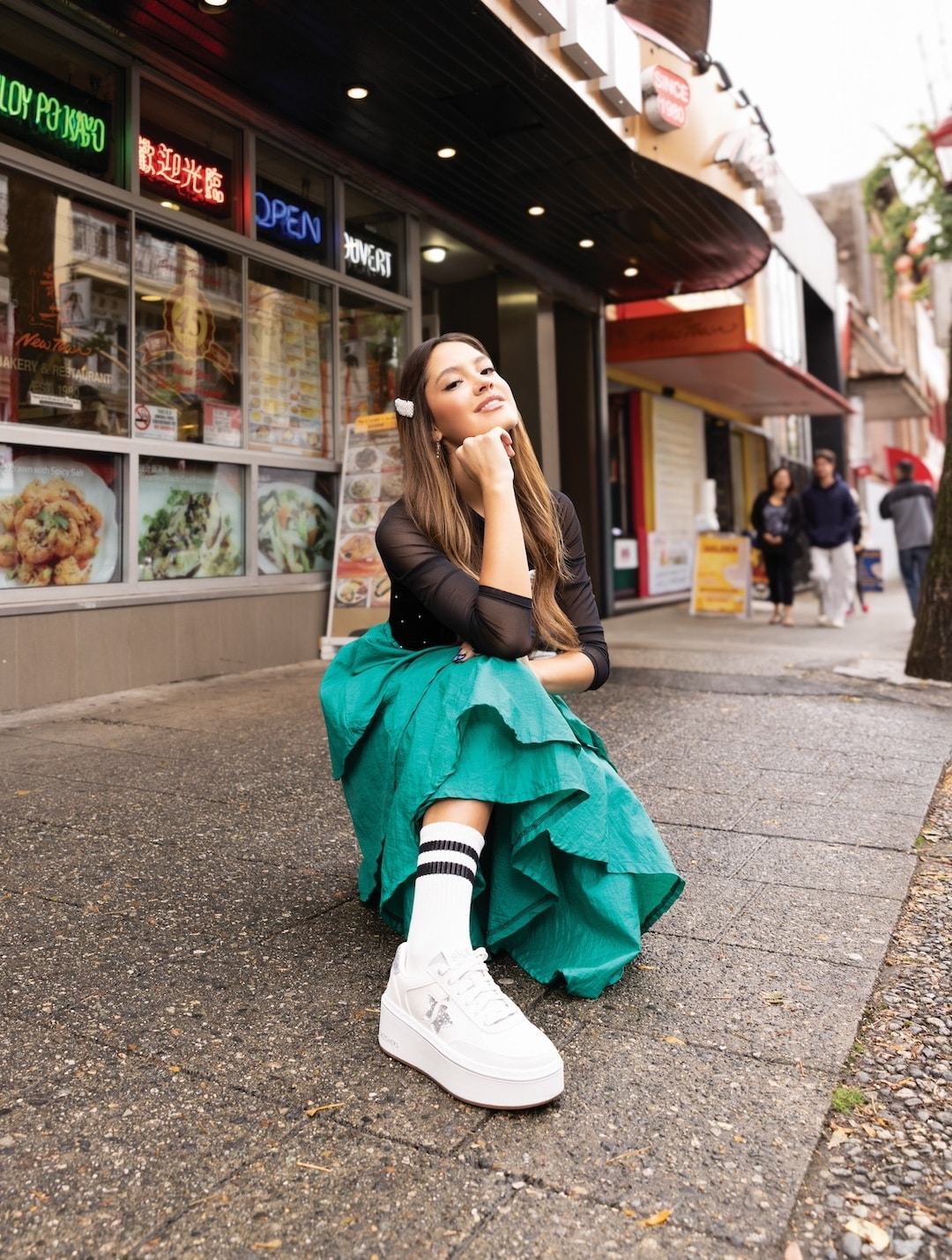 A young woman in a green skirt, black top, and white platform sneakers poses confidently on a city sidewalk in front of colorful 2026 restaurant signs, with pedestrians walking in the background.