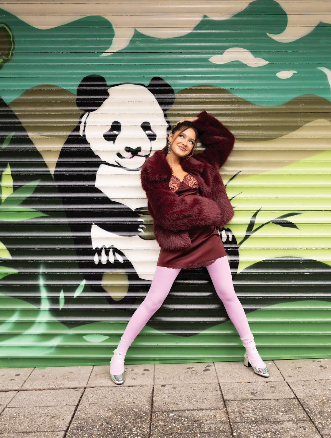 Liamani Segura poses playfully in a burgundy jacket, short dress, and pink tights in front of a mural featuring a smiling panda on a green and teal background, bringing vibrant 2026 fashion vibes to the scene.