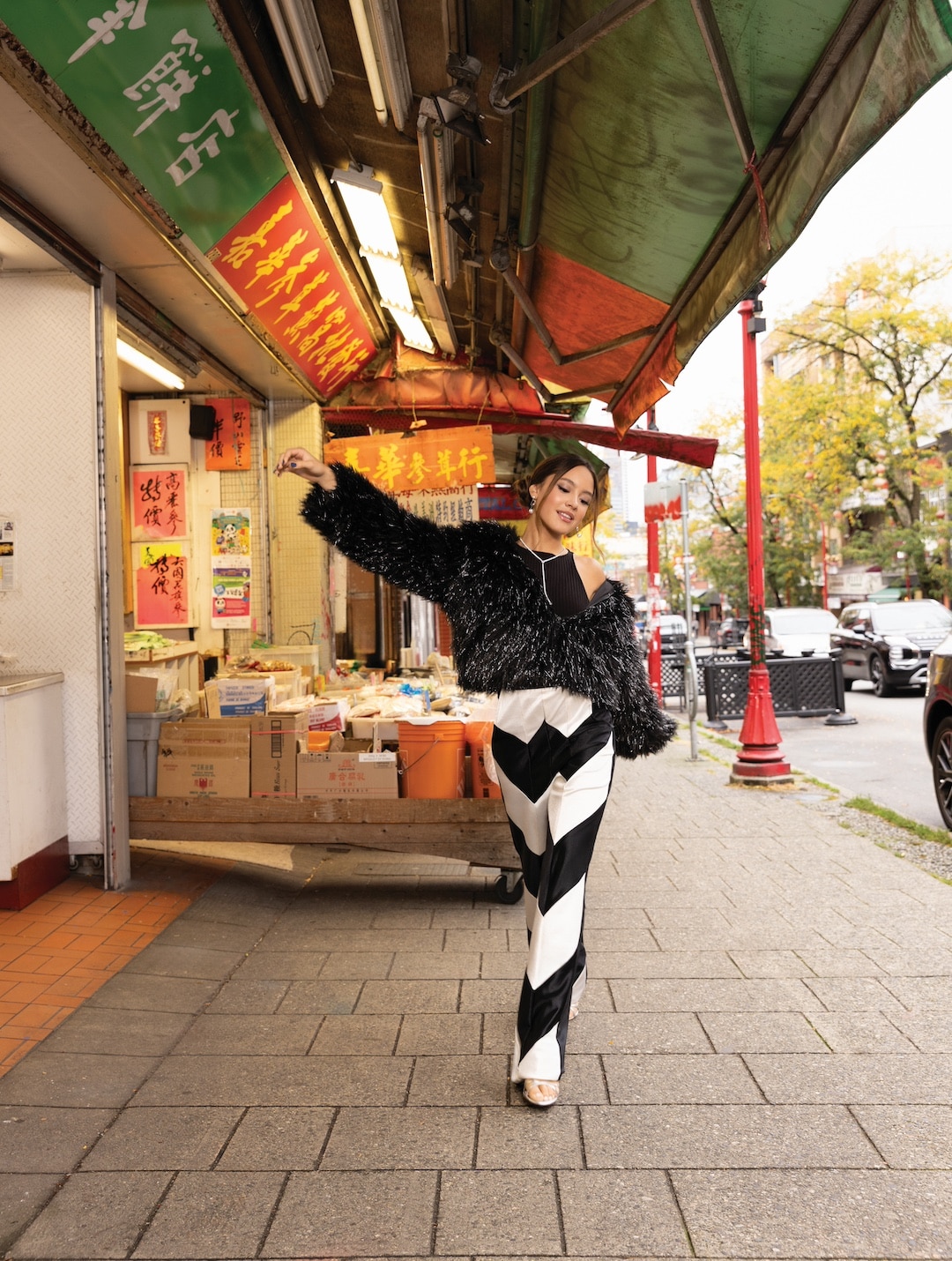 Liamani Segura poses confidently in a fuzzy black sweater and bold black-and-white striped pants on a city sidewalk near a market with red and green awnings and Asian signage, embodying chic urban style in 2026.