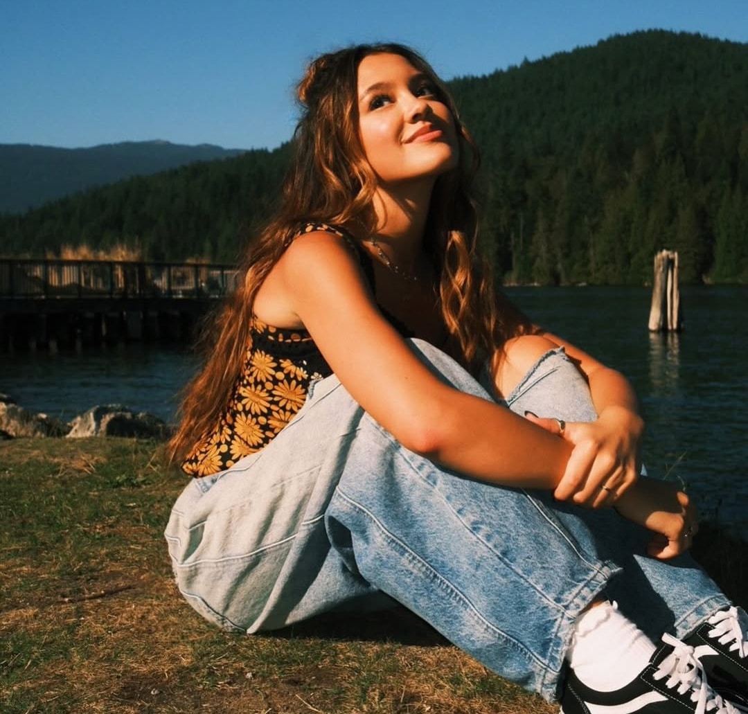 A young woman with long hair sits cross-legged on grass by a lake, wearing a floral top, jeans, and sneakers. She looks up and smiles, with green forested hills and blue sky in the background—like a scene from Liamani Segura’s 2026 tour.