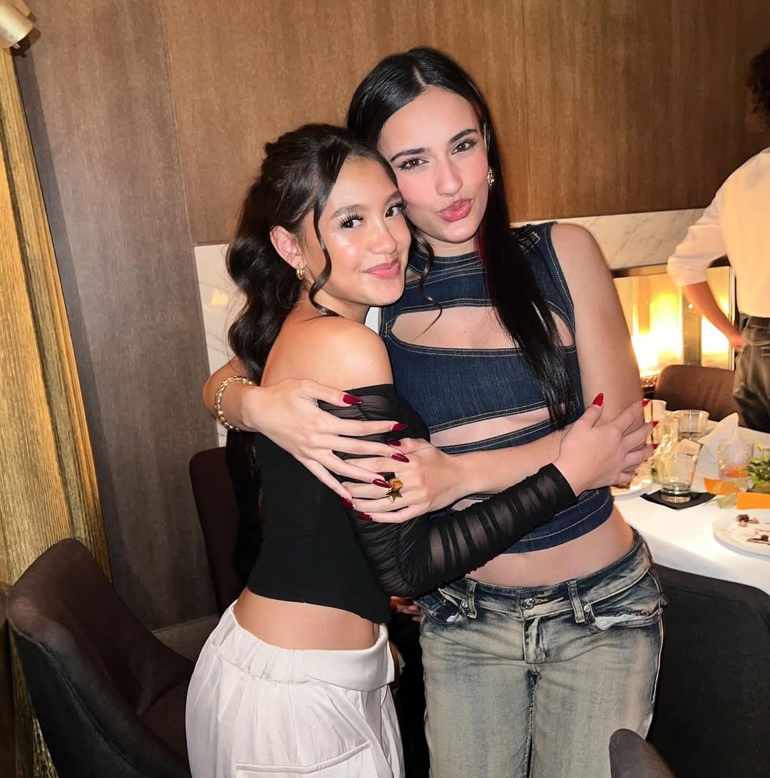 Two young women, including Liamani Segura, are standing close together and smiling at the camera while hugging in a warmly lit restaurant. There is food and drink on the table behind them, capturing a joyful 2026 evening.