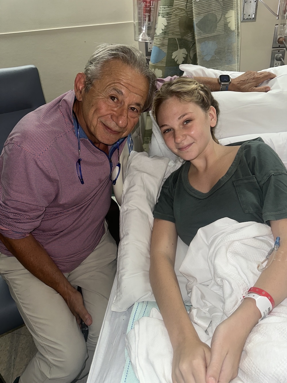 An older man smiles and leans next to a young girl lying in a hospital bed. Despite her period cramps, the girl, with an IV in her arm and a hospital band on her wrist, smiles gently at the camera.