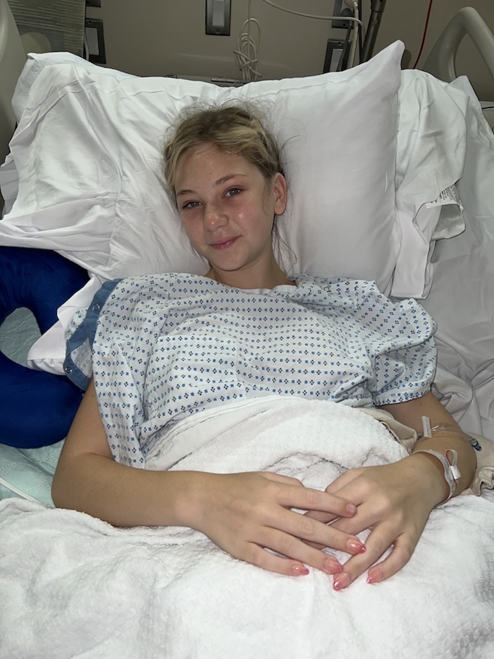 A young girl with blonde hair lies in a hospital bed, wearing a hospital gown and covered with a white blanket. She has a slight smile and an IV attached to her arm. Pillows are propped behind her for support.