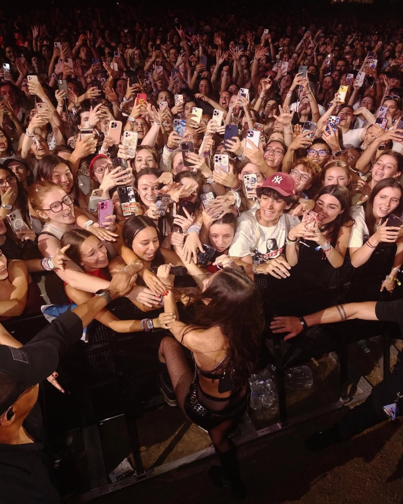 A singer kneels at the edge of a stage, reaching out to excited fans in a large, packed crowd. Many fans are reaching toward her and holding up phones to take photos or videos.