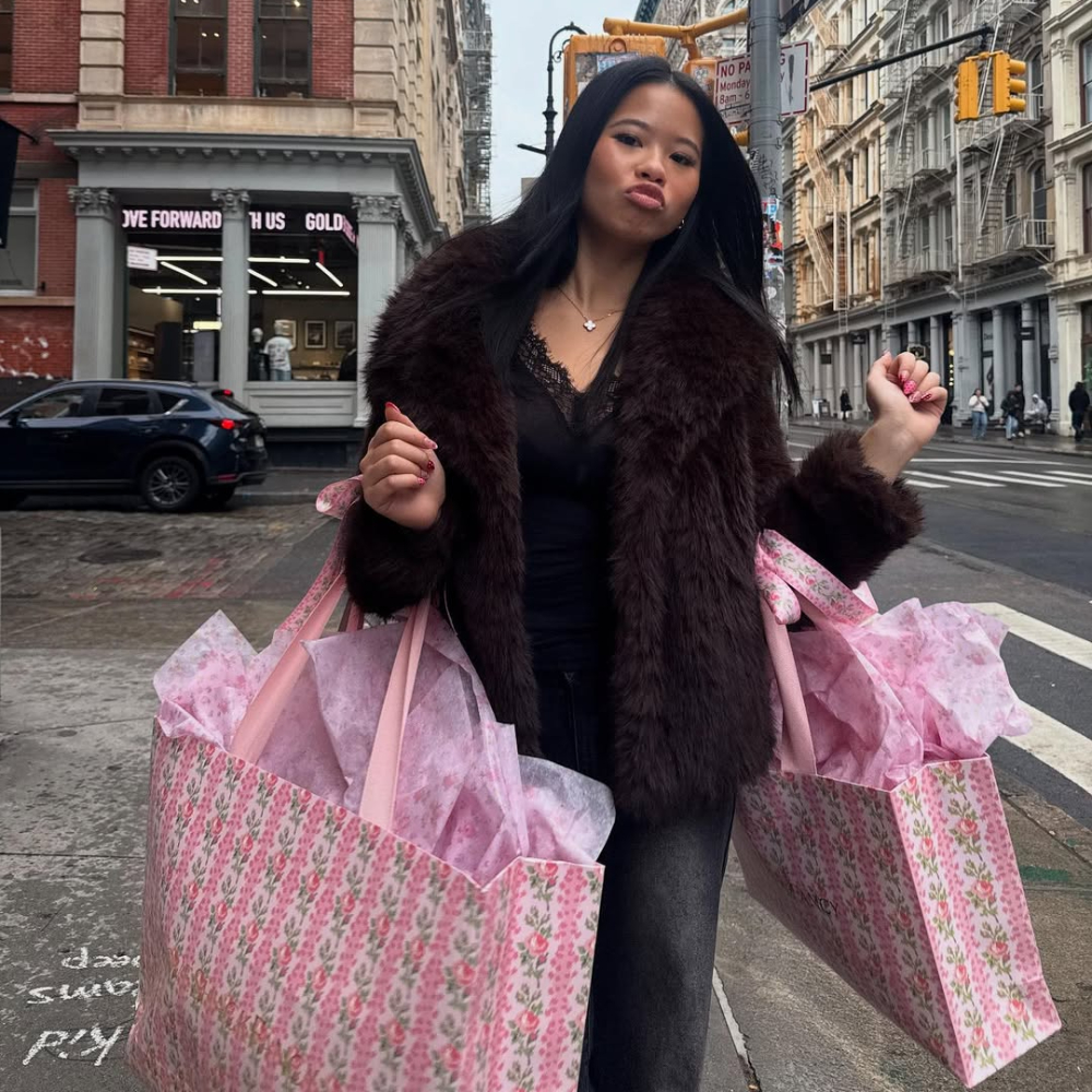 A woman wearing a dark fur coat and black top stands on a city street, holding two large pink floral shopping bags filled with tissue paper—showing up for your bestie in true friendship style. She pouts, her long dark hair adding to her chic look.