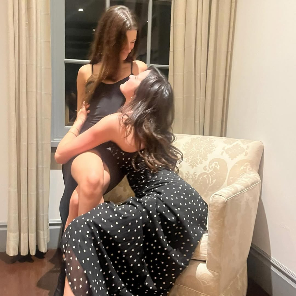 Two women in black dresses, one with polka dots, pose closely together on an armchair in a softly lit room with cream-colored curtains and walls—capturing a heartfelt moment of supporting friends and understanding each other’s love languages.