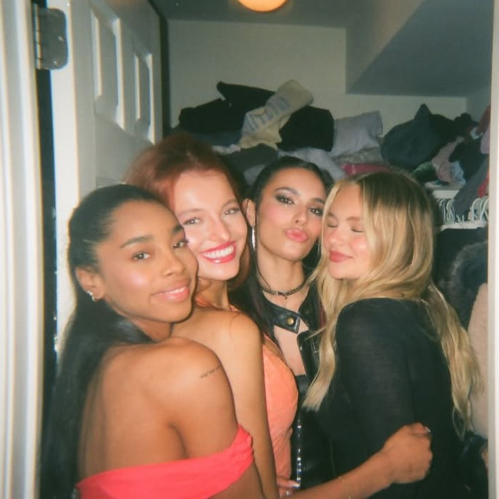 Four young women are posing closely together and smiling in a small, cluttered room with clothes in the background. Three look at the camera while one, eyes closed, hugs her bestie—showing that hugs are her love language.