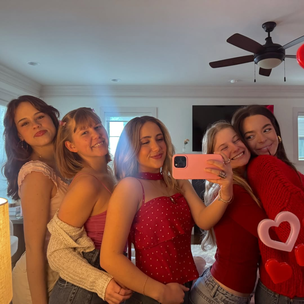 Five young women smiling and posing for a mirror selfie in a brightly lit room. One bestie holds a pink phone, and several wear red or pink tops. Heart decorations on the mirror reflect their love language of showing up for each other.