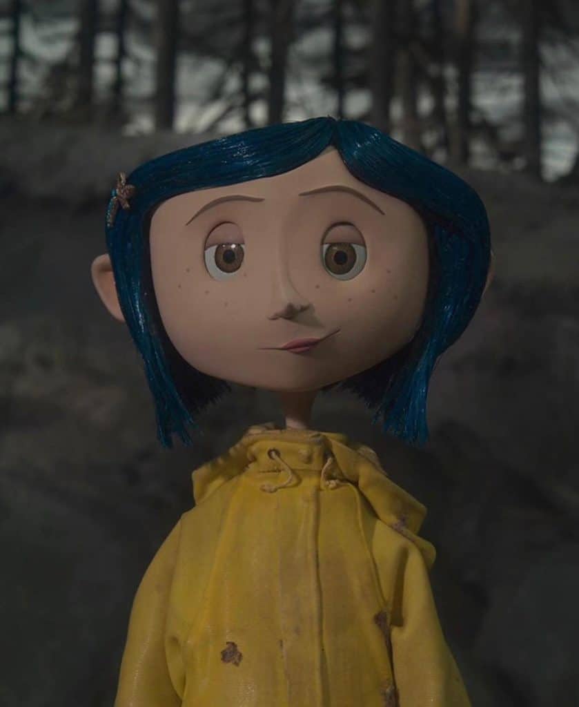 A stop-motion animated girl with blue hair and a yellow raincoat stands outdoors in front of a dark, blurred forest background. She has wide eyes and a small, closed-mouth smile.