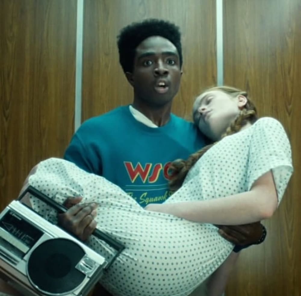 A shocked young man, resembling a Stranger Things character, holds an unconscious girl in a hospital gown. He carries a large portable cassette player as they stand before wooden elevator doors.