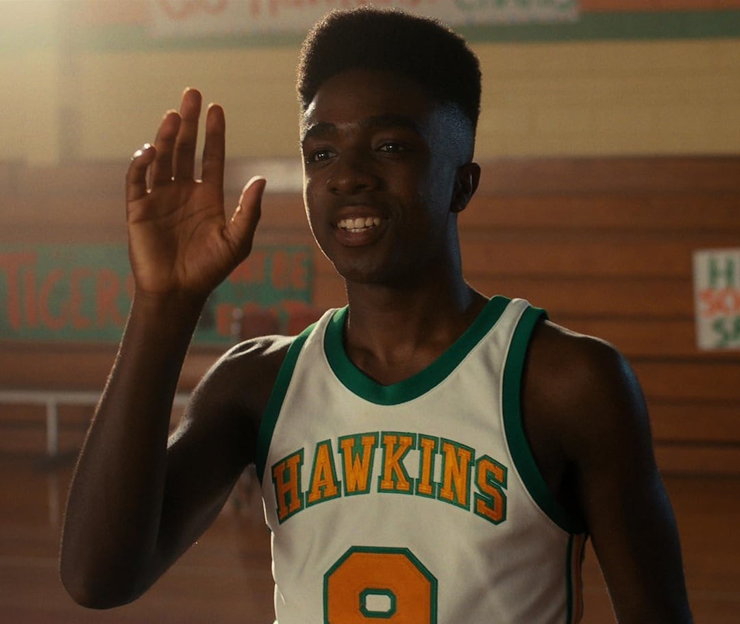A young man wearing a white and green basketball jersey with HAWKINS and the number 8 raises his hand in a gymnasium with wooden bleachers, channeling Conrad Fisher's confidence on Valentine's Day.