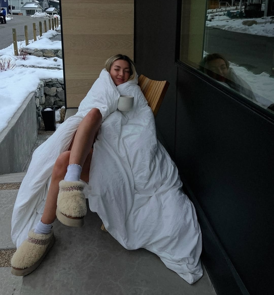 A person wrapped in a white comforter sits on a chair outside, holding a mug. They wear cozy slippers and socks, embracing the chill while snow covers the ground—an ideal scene for reflecting on what to expect during different menstrual cycle phases.