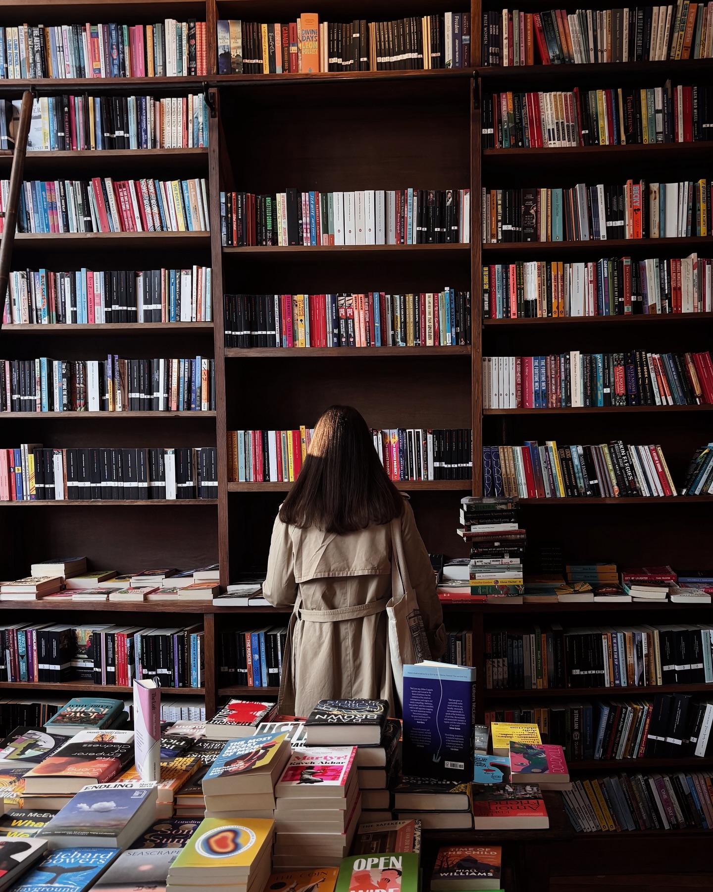 A person in a beige trench coat stands with their back to the camera, browsing tall bookshelves filled with classic books in a bookstore. Books are stacked on tables in the foreground.