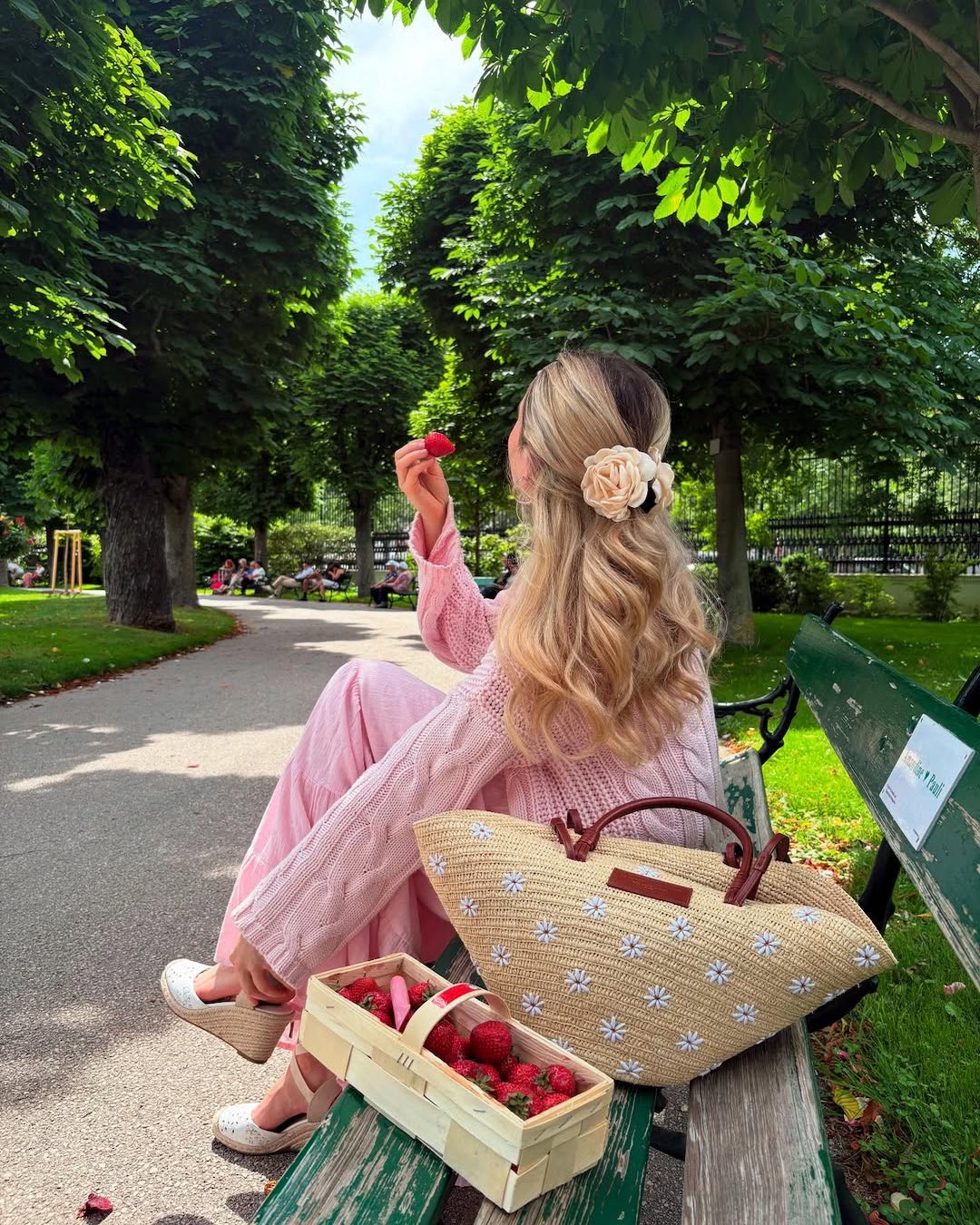 A woman with long, wavy blonde hair sits on a park bench, holding a strawberry. She wears a pink dress and white shoes, with a basket of strawberries and a straw tote bag beside her. The park is lush and green.