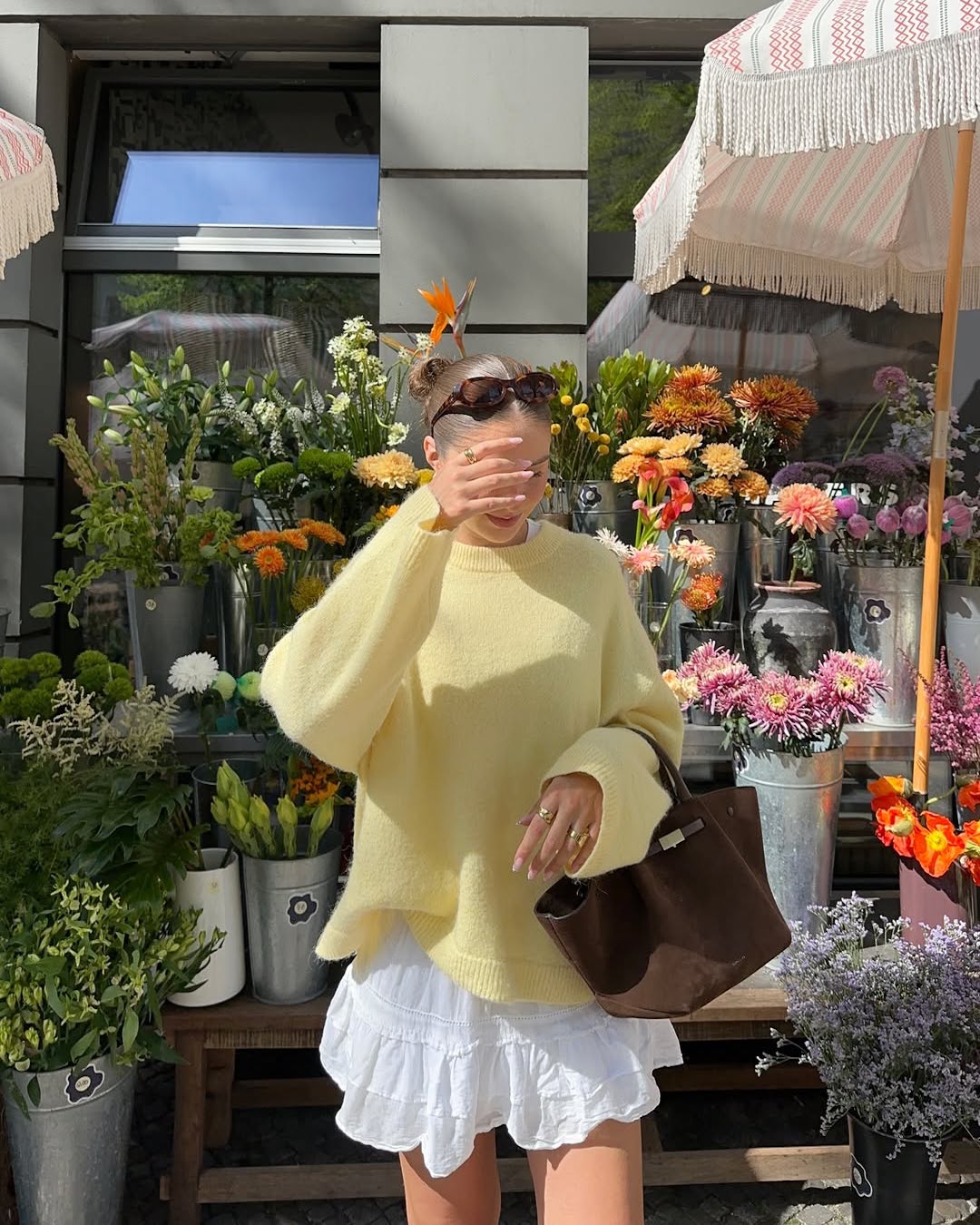 A person in a yellow sweater and white ruffled skirt stands in front of a flower shop, holding a brown bag and covering their face from the sun with their hand. Buckets of colorful flowers are in the background.