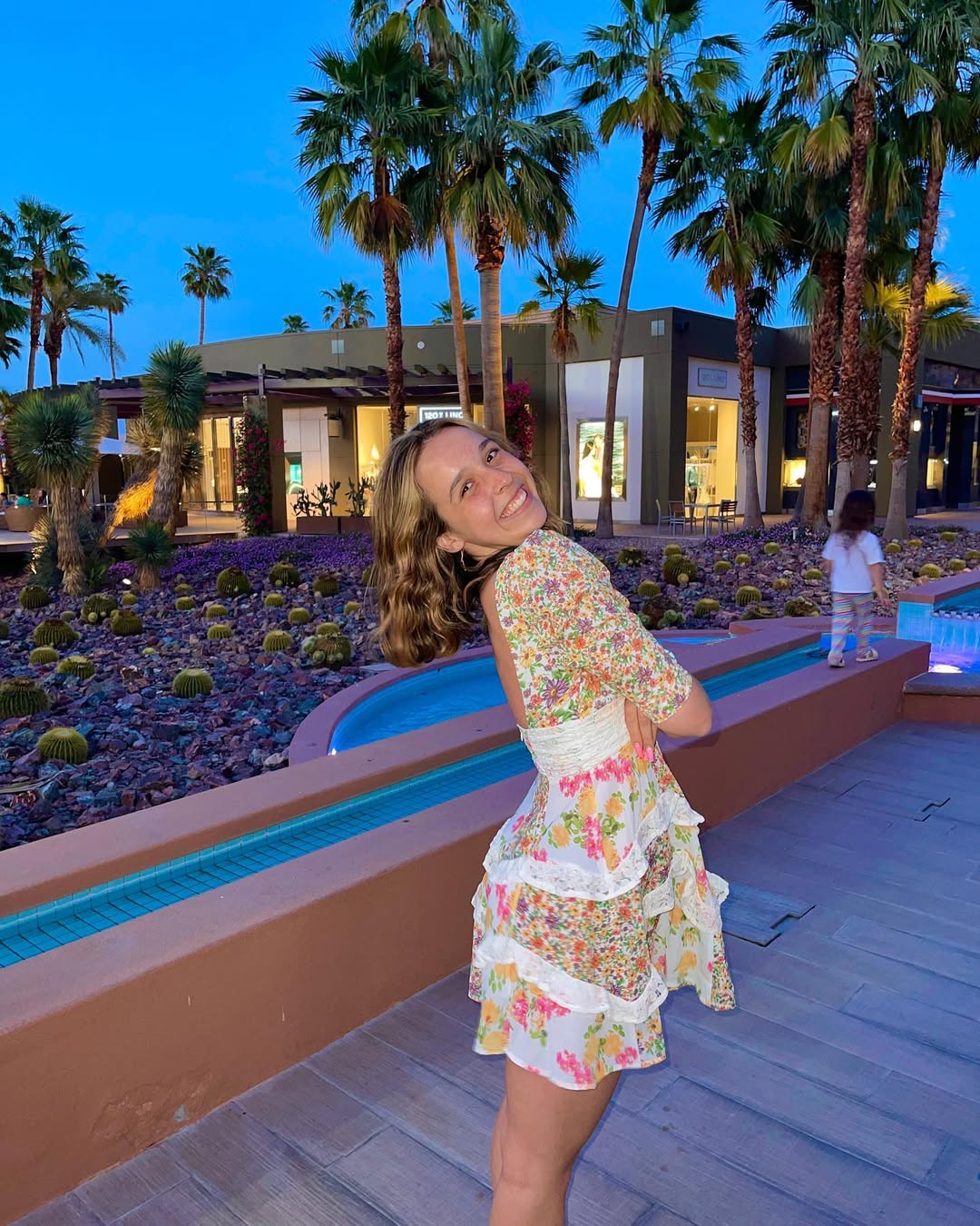 A smiling woman in a floral dress stands outdoors at dusk, posing near a curved railing. Palm trees and shops are visible in the background, and a young child walks nearby.