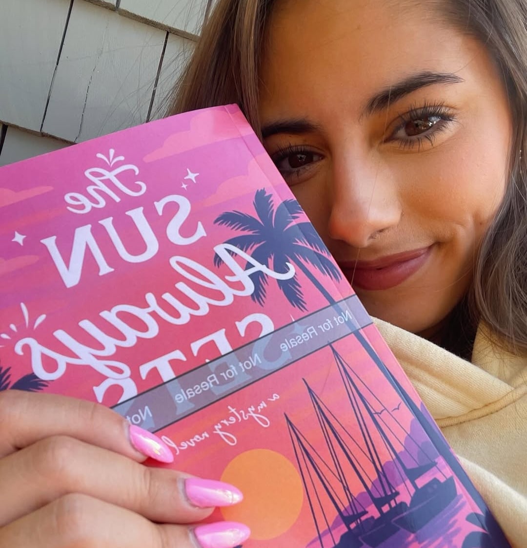 A young woman with long brown hair and pink nails smiles while holding a book titled The Sun Always Sets, its pink and purple cover featuring palm trees and a sunset—proof that anyone can Become the Author of their own success story.
