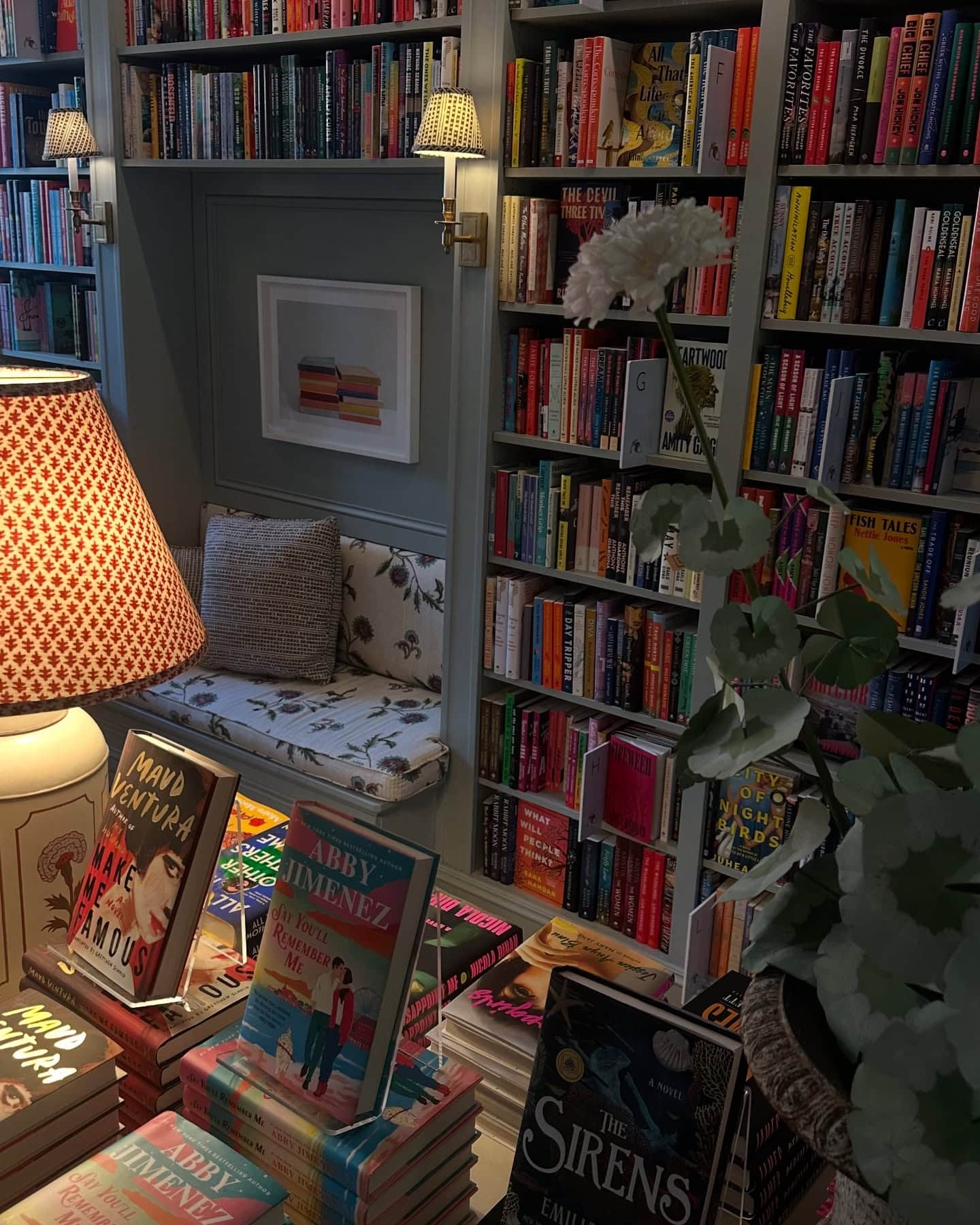 A cozy bookstore with shelves full of colorful books, a cushioned bench under framed art, a lamp with a patterned shade, and a flower in a pot next to a display table with stacked novels.