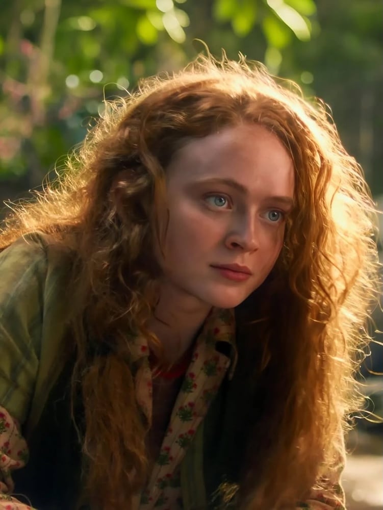 A young woman with long, curly red hair and blue eyes looks intently forward. She is outdoors, surrounded by greenery, and wearing a patterned shirt with a plaid jacket. Sunlight filters through the background.