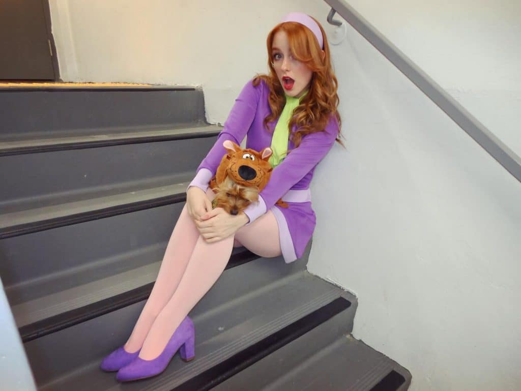 A woman in a purple dress, headband, and heels, dressed as Daphne from Scooby-Doo, sits on stairs holding a small Scooby-Doo plush toy, with a surprised expression.