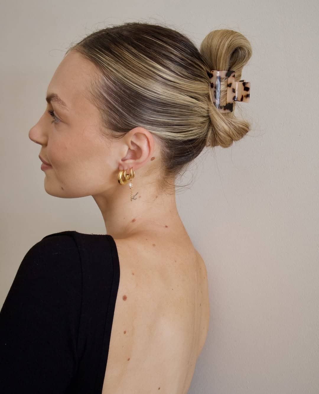 A woman with light skin and blonde hair pulled into a bun secured with a stylish claw clip, wearing gold hoop earrings and a black, open-back top, is pictured in profile against a plain light background.