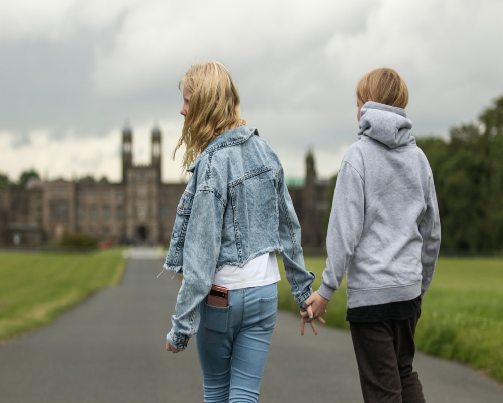 Two young people, likely BFFs, hold hands as they walk down a paved path toward a large historic building. One wears denim, the other a grey hoodie. It’s cloudy—perhaps theyre talking about going to a different school soon.